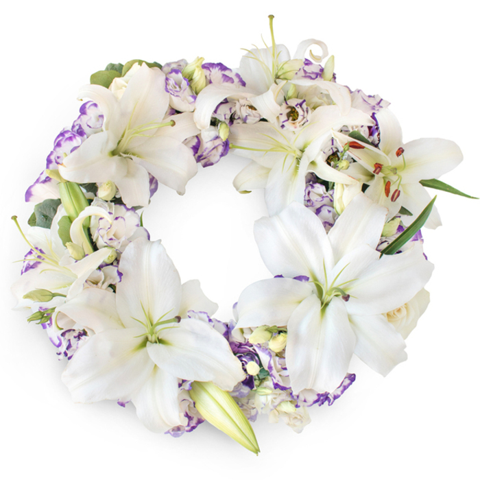 A meticulously arranged circular wreath of sympathy flowers is depicted floating against a pure white background, its lush form hiding the underlying frame beneath a seamless layer of blossoms. Striking white Oriental lilies are the centrepiece of the design, spaced thoughtfully around the circle so their graceful, star-shaped blooms appear to almost overlap. The petals are broad and satiny, some lying open and flat, others arching and curling at the tips. At the heart of each flower, a soft green throat blends into long, slender stamens bearing golden-brown pollen, providing a warm accent amid the cool whites. Nestled between the lilies are generous sprays of lisianthus blossoms, their petals delicately ruffled and tipped with shades of lilac, lavender, and gentle purple. These purple-edged flowers weave through the wreath like a soft ribbon of colour, adding visual rhythm and depth without disturbing the overall calm. Here and there, small, creamy rosebuds and double lisianthus blooms add an extra layer of texture and tenderness, evoking the feeling of a carefully tended garden. Slender leaves and eucalyptus-like sprigs in rich and silvery greens surface just enough to give structural support and a whisper of nature's wildness. The lighting is clear and even, accentuating every subtle contour while maintaining a respectful, tranquil mood. This is the kind of wreath a family in Mill Hill might choose for a service at a nearby church, or for a memorial gathering at a local venue. Lovingly crafted by Flowers Mill Hill, it conveys elegance and serenity, honouring a life with understated beauty and a sense of continuing love encircling the memory of the departed.