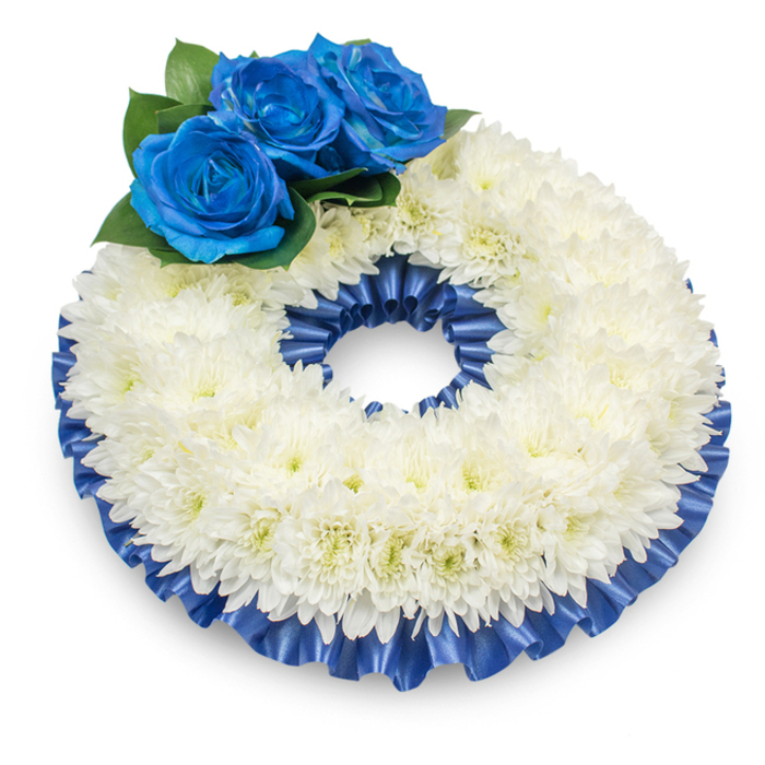Captured from slightly above on a plain white surface, this traditional circular funeral wreath feels both intimate and formally composed. The main body is densely arranged with immaculate white chrysanthemums whose innumerable feathery petals build a soft, pillowed circumference that symbolises purity and remembrance; a very faint creamy centre peeks through some blooms, lending a whisper of warmth to the cool white. At the wreath's upper-left sector, three electric-blue roses are clustered as a deliberate focal point-their velvety, saturated petals unfurl with precise, sculpted edges and are set amidst deep green foliage that accentuates their intensity. A lustrous cobalt satin ribbon, pleated and gently ruffled, frames the outer and inner perimeters, its glossy surface catching highlights and reinforcing the wreath's circular geometry. The subtle shadow the arrangement casts on the white background gives it a sense of weight and presence without heaviness, inviting close inspection of texture and finish. Speaking as a florist who makes many memorial pieces for families around Mill Hill Broadway, Bittacy Rise and nearby chapels, I can say the restraint and balance here are intentional: the white chrysanthemums provide a communal language of condolence while the blue roses allow a personal, signature note. The overall impression is of calm dignity-textural yet neat, classic yet personal-appropriate for a service at a local church or cemetery, carried in with quiet respect and intended to console while marking the lasting memory of the departed.