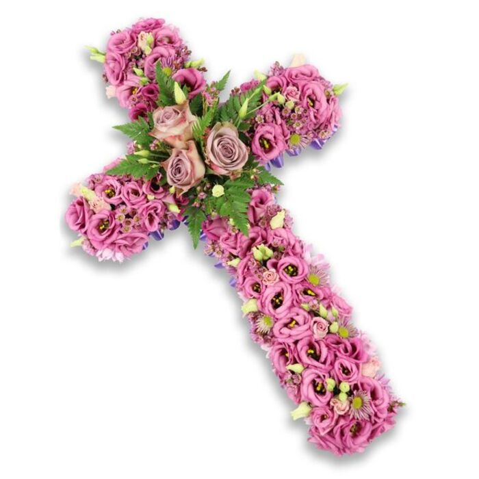 This photograph presents an exquisite floral cross tribute, centrally framed against a stark white background that amplifies the colours and contours of each bloom. The cross is thoroughly enrobed in a rich tapestry of flowers, primarily soft pink and mauve lisianthus whose ruffled petals create a gentle, cloud-like texture from top to base. Their tones shift delicately from pastel blush to more saturated rose and lilac, with some flowers revealing darker centres that lend a slightly mottled, painterly effect across the surface. At the crossing point of the arms, three large roses become the deliberate focal feature, opening fully in shades of dusty rose and muted mauve. Their classic, spiralled petals add a sense of poise and tradition, suggesting enduring love and remembrance. These central roses are cushioned by arcs of fresh green fern fronds and small, bright leaves, the foliage slipping between the petals to add structure and an organic rhythm to the piece. Nestled amid the lisianthus along the arms are tiny pink and purple fillers, specks of pale cream blossoms, and budding green tips that evoke the quiet promise of new life. Around the perimeter of the cross, slender suggestions of darker purple ribbon or fabric can be seen beneath the flowers, neatly defining the silhouette and reinforcing the strength of the Christian symbol. The entire arrangement conveys a mood of serene reflection and heartfelt respect, as though it has been thoughtfully created by Flowers Mill Hill for a local family saying goodbye at a church near The Ridgeway or during a service close to Mill Hill's leafy lanes, offering a dignified floral tribute that speaks gently of faith and memory.