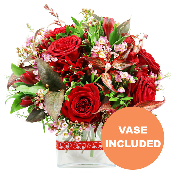 Set against a bright white background, this opulent arrangement of red and green feels like a romantic centerpiece waiting to be delivered to a home in Mill Hill. At its core, velvety red roses command attention, their petals rich and plush, with some buds tightly closed and others unfolding to reveal intricate spirals of crimson. Interwoven with these roses are clusters of smaller deep red blooms, reminiscent of alstroemeria or similarly textured flowers, adding dimension and a sense of fullness to the composition. Fine sprays of tiny pale pink and white filler flowers, much like waxflower or limonium, drift between the larger stems, providing a gentle, airy contrast that lightens the drama of the reds. Surrounding the flowers is an abundance of fresh greenery: large, glossy leaves anchor the design, while more delicate green sprigs offer movement and softness, calling to mind the lush foliage found in the gardens around Mill Hill. Among the greens, distinctive burgundy and reddish-brown leaves are strategically placed, some with subtle veining that echoes the tones of the roses and adds a layer of sophisticated depth. The bouquet is shaped into an elegant, rounded dome, meticulously crafted so that the arrangement appears full and balanced from every side, as though carefully composed for a romantic dinner or anniversary celebration. The stems are neatly gathered into a sleek, clear glass vase with a rectangular base and gently rounded corners, allowing the clean lines of the container and the stems to complement the blooms. Around the vase is a charming red ribbon decorated with a continuous pattern of white hearts, reinforcing the bouquet's message of love and affection. In the bottom right corner of the image, a bold circular orange overlay with crisp white uppercase text clearly reads VASE INCLUDED, highlighting that this striking, gift-ready bouquet arrives complete for Valentine's Day, anniversaries, or romantic gestures across Mill Hill.
