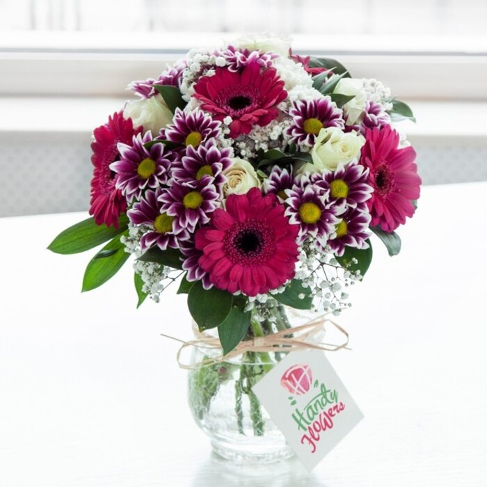 A striking mixed flower arrangement brimming with colour and texture stands in the centre of the frame, cradled by a clear, subtly textured glass vase that catches the light in fine, glimmering ripples. The bouquet is composed around several large gerbera daisies in vivid fuchsia and magenta shades, their dark, velvety centres providing a dramatic contrast to the bright petals. Surrounding these focal blooms are generous clusters of chrysanthemum-style flowers, each floret a rich purple at the edges and gently fading to clean white near the centre, where warm yellow discs echo the glow of late-morning sun over Mill Hill's tree-lined streets. Creamy white roses and compact rose sprays are scattered thoughtfully throughout the design, some in full, layered bloom, others still in tight bud form, adding a quiet sophistication and a sense of unfolding moments. Delicate trails of white gypsophila thread in and out of the arrangement like soft mist, filling spaces between the larger blooms and giving the bouquet an airy, almost cloud-like halo. Deep green foliage peeks between petals and fans out subtly around the base, providing structure, depth, and the grounding scent of fresh leaves. The vase itself has a charming, jar-like silhouette and is finished with a rustic loop of natural twine tied neatly at the neck. From this twine hangs a small, crisp white tag, printed with a pink rose emblem and flowing script, suggesting a personalised note accompanying a special delivery. Bathed in diffused, almost studio-like light against a clean white backdrop, the flowers appear exceptionally fresh and vibrant, evoking the feeling of a thoughtfully chosen gift arriving at a home near Mill Hill Broadway or along Daws Lane to celebrate a birthday, an anniversary, or simply to brighten a calm weekend morning.