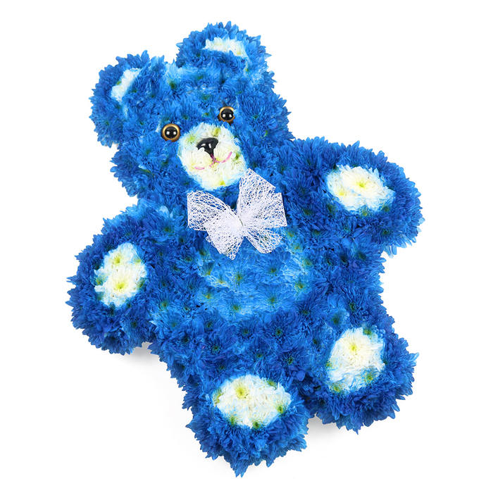 This image shows a three-dimensional teddy bear made entirely of flowers, resting against a clear white background in a pose that suggests peaceful sleep, much like a cherished toy laid gently down in a Mill Hill bedroom or chapel. The bear's form is defined by hundreds of royal blue blossoms, likely chrysanthemums or similar button flowers, packed so tightly that every inch of the body appears covered in soft, petal-based fur. The blue hue is intense and jewel-like, deepening into shadows along the underside of the arms, legs, and chin, while catching light on the rounded belly and ears. Accents of creamy white flowers, with subtle buttery centres, shape the rounded muzzle, the circular inner ears, and the four oval pads on each paw, giving the figure clear definition and a sweet, childlike character. Two glossy circular eyes, dark brown with black pupils, are embedded neatly within the blue petals, gazing outward with a quiet, steady expression. Below them sits a small black triangular nose, with a delicately curved black line beneath that forms a modest, comforting smile. Around the neck, a fine white mesh bow is tied in soft, semi-transparent loops that fan out lightly against the blue, adding an airy, celebratory touch. The bear lies at a gentle angle, which creates a relaxed, natural composition rather than a stiff, upright pose. One can imagine this floral teddy placed near a window in a Mill Hill home overlooking the green spaces or in a chapel by Mill Hill Broadway, its rich colours and intricate craftsmanship offering solace and remembrance at a funeral, baby memorial, or as a tender sympathy tribute for a young life.