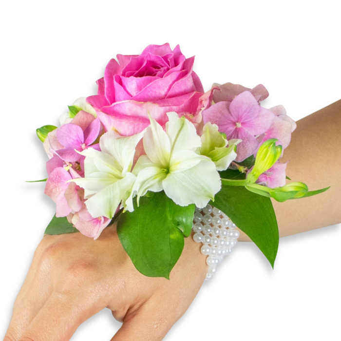 In clear, even lighting against a white studio background, a corsage is displayed on a left hand and forearm, showing the kind of detailed workmanship we create at Flowers Mill Hill for local ceremonies and celebrations. The centrepiece is a bold deep fuchsia-pink rose, opened fully so each layered petal radiates outward with a plush, velvety texture. Surrounding the rose are clusters of soft, blush-pink hydrangea florets that give the arrangement a rounded, romantic fullness and a gentle translucence at their edges. Interspersed are white alstroemeria (Peruvian lilies), each bearing four to six graceful petals subtly streaked with pale green toward their throats, offering crisp highlights against the deeper hues. On the right side of the composition a grouping of pale green alstroemeria buds and a few smaller, nearly closed pink buds introduces a sense of growth and anticipation. Broad, rich emerald leaves cradle the blossoms, their glossy surfaces and pronounced veins creating depth and contrast. The corsage is secured by a multi-strand bracelet of small, luminous white pearls that wrap the wrist and trail slightly across the back of the hand, lending a refined, heirloom touch. The tactile contrasts - the velvet of the rose, the soft papery hydrangea, the smooth pearls and cool leaves - are evident even in this portrait-style image. Ideal for prom, weddings, or graduation moments across Mill Hill Broadway and nearby streets, this wearable bouquet reflects local floristry skill, attention to proportion, and the kind of freshness we bring to same-day orders and bespoke requests within NW7.