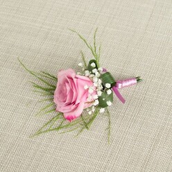 Set against a pale, textured linen fabric that recalls the relaxed elegance of a Mill Hill dining room dressed for a celebration, a single pink rose boutonniere is captured from a slightly overhead perspective. The rose itself is in full, confident bloom, with layers of velvety petals spiralling outward from a softly cupped centre. Its colour moves gently from a tender blush at the heart to a more vibrant, romantic pink at the petal tips, giving the flower a sense of depth and freshness, as though it has just been prepared in a nearby Mill Hill florist's studio. Nestled closely around the base of the rose are clusters of delicate white baby's breath, their tiny rounded blossoms forming a mist-like halo that lightens and frames the stronger colour of the central flower. Fine, wispy strands of light green foliage arc out behind the rose, creating a subtle fan of texture and movement, while a single, broader deep green leaf is wrapped and folded to cradle the stems, adding structure and a clean, tailored edge. At the base of the boutonniere, the stems are neatly bound with smooth pink satin ribbon, tied into a simple, elegant bow with short trailing ends that catch the light. The soft, natural lighting emphasises the gentle curve of each petal, the satin sheen of the ribbon, and the woven pattern of the beige background, without casting harsh shadows. This thoughtfully composed image conveys the calm joy of a wedding morning or school prom near Mill Hill Broadway or Bittacy Hill, and shows a classic rose boutonniere ready to be pinned securely to a lapel for a day filled with romance, photographs, and shared memories.
