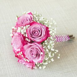 Illuminated by soft daylight that hints at Mill Hill's gentle hillside mornings, this bridal bouquet is a celebration of colour, texture, and timeless romance. Its heart is formed by five vividly bi-coloured roses-each bloom glowing from within as lavender and light purple petals give way to saturated, jewel-bright fuchsia at the tips. Between the unfurling roses, wisps of white baby's breath impart an airy, cloudlike delicacy. The pleasing contrast between velvet-rich petals and featherweight gypsophila is beautifully balanced, shown to advantage atop a creamy, off-white linen that adds warmth and textural interest. The short stems are meticulously bound in sumptuous plum ribbon, accented with parallel spirals of lustrous white pearls, recalling both sophistication and the cherished wedding traditions of Mill Hill's vibrant community. Beneath this decorative finish, the freshly snipped green stems whisper of gardens just steps from Mill Hill Park. Crafted by local artisans, this bouquet expresses both tenderness and contemporary flair-an intimate floral companion for a wedding at a neighbourhood venue, a special anniversary on The Ridgeway, or a celebration amongst friends in a mill house garden. Through its thoughtful arrangement and radiant palette, the bouquet mirrors the joyful anticipation and cherished memories woven into every Mill Hill ceremony.