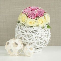 On a smooth white tabletop, a refined bouquet of roses glows softly, as if prepared for a wedding breakfast at a stylish venue near Mill Hill Broadway. Around a dozen roses form a perfectly rounded dome, carefully placed so that creamy butter-yellow blooms sit harmoniously alongside cool lavender-pink or fuchsia roses. The petals show a beautiful variety of shapes and stages, from tight, sculpted curls at the centre of younger blooms to broader, gently unfurling layers in the more open flowers. Between them, feathery sprays of white baby's breath weave in and out, dotting the arrangement with clusters of tiny blossoms that add lightness and a sense of airy romance. The bouquet is set into a striking, spherical vase constructed from slender, white-painted wicker or twig-like strands. This open-weave design creates a delicate cage around the blooms, casting fine shadows on the white surface below and offering glimpses of the stems and inner support. To the left of the central piece, two decorative orbs made from pale natural rattan echo the same woven motif, their different sizes lending balance and subtle rhythm to the display. The background is a soft, neutral beige or taupe wall with a subtle, fabric-like texture, keeping the focus firmly on the flowers and their intricate detailing. The lighting is gentle and even, without harsh contrast, evoking a calm, welcoming mood that would suit both intimate weddings and relaxed family gatherings across Mill Hill. This arrangement feels ideal for describing elegant table flowers delivered locally, adding a touch of understated luxury to homes and celebrations in the area.