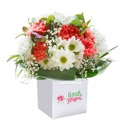 Celebrating the artistry and garden-inspired excellence of Mill Hill's local florists, this bouquet gleams with lively colour and meticulous composition. Set proudly in a stark white, square gift bag topped with wide ribbon handles for easy carrying, its rounded form promises abundance and cheer-much like a thoughtful surprise waiting on a doorstep along Mill Hill East. The heart of the arrangement features myriad coral and soft peach carnations, their ruffled, textured petals spilling generously in layered waves. Pure white daisies, bright with zesty green-yellow centers, peek from between the carnations, infusing the bouquet with a playful, sunny mood that recalls the open sky over Mill Hill Park. Subtle, airy clusters of white baby's breath create gentle movement and balance alongside bold foliage, while a handful of small, closed buds tinged with purple on the left add a sense of mystery and depth. Around the base, shiny, deep green leaves wrap the flowers in a natural, structured embrace, lending stability and richness to the display. Careful lighting brings out the vibrant hues and contrasts, revealing a bouquet ready for any joyful occasion-a birthday along Salcombe Gardens, an anniversary, or an act of gratitude. This arrangement, available for delivery throughout Mill Hill, demonstrates the florist's skillful touch, creating a lasting impression of freshness, warmth, and thoughtfulness.