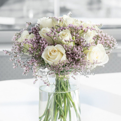 In this softly focused photograph, a refined sympathy bouquet stands tall in a clear glass vase, echoing the gentle character of Mill Hill's leafy streets and calm residential spaces. The arrangement is composed predominantly of creamy white roses, each one fully open and at its most expressive stage, revealing intricately layered petals that form a classic spiral at the core. Their texture appears velvety and cool, the white softened by a hint of ivory that adds warmth and depth, as if to mirror the kindness behind a condolence gesture. Threaded delicately between the roses are sprays of fine-textured filler flowers in shades of lavender, pale purple, and the faintest blush pink. These tiny blooms gather like a light mist, giving the bouquet a soft halo effect that makes the roses appear to float in a haze of pastel colour. All the stems are arranged in a tall cylindrical vase of perfectly clear glass, filled with clean water in which the green stems are clearly visible, some slightly angled, others straight, resembling gentle lines drawn downward. The vase rests upon a smooth white surface that reflects soft natural light, while the background is intentionally blurred into tones of white and soft grey, suggesting a quiet, modern interior somewhere near Mill Hill Broadway or along the quiet avenues leading toward Bittacy Hill. The overall feeling is one of tranquillity and restraint, with every detail contributing to an atmosphere of respect and peaceful remembrance. This is the kind of bouquet that a local Mill Hill florist might prepare for a memorial service or to send as a thoughtful message of sympathy, letting the flowers speak with quiet grace.
