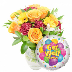 Bathed in soft daylight, this uplifting 'Get Well Soon' arrangement reads like a note of genuine encouragement from a neighbour. A lush bouquet, slightly left of centre, is gathered in a clear glass vase that reveals the straight green stems and fresh water below; glossy foliage fans out to give structure and a lively green backdrop. The floral story begins with large, warm orange and deep yellow roses whose velvety petals open outward in gentle spirals, exuding a soft, citrus-rose scent that feels calming. Bright yellow gerbera daisies bring sunny faces and instant cheer, while maroon-to-burgundy carnations lend a richly textured, cushion-like depth. Sprays of white alstroemeria punctuate the grouping with delicate flecks of light, creating graceful moments of contrast. Nestled to the lower right, a silver Mylar balloon with a metallic gloss overlaps the bouquet slightly, its centre illustrating a cluster of colourful, patterned balloons in sky blue, purple, lime green, pink and yellow. The message "Get Well Soon" appears in bold yellow-and-blue letters with a slight drop shadow, and the balloon's border is dotted with playful starbursts; a tiny "KALEIDOSCOPE" logo is visible at the bottom edge. Prepared by Flowers Mill Hill with an experienced florist's eye, this pairing is ideal for delivering cheer to someone convalescing at home or receiving care near The Ridgeway or Mill Hill Village, and is equally suited for same-day local delivery around Mill Hill East and nearby streets.