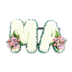 In this serene funeral flower arrangement, the letters MA stand side by side, forming a classic tribute that many families choose to honour a much-loved Mum, Mama, or grandmother. Both letters are crafted on a sturdy base and then lavishly covered with flawless white chrysanthemums. Each bloom is placed closely against the next, creating a dense, gently undulating surface that looks almost like freshly fallen snow captured in floral form. The chrysanthemums' fine, intricate petals lend a velvety softness and symbolize purity and remembrance. Running neatly around the outer edge of each letter is a dark green border, giving the tribute a crisp, tailored outline. This edging, made from ribbon or carefully arranged foliage, defines the curves and angles of the M and the A, adding an elegant finish that would look fitting on a hearse travelling through Mill Hill, from quiet residential streets to nearby churches or Hendon Cemetery. At the base of the left leg of the M and the right leg of the A, gentle sprays of colour soften the whiteness. Clusters of soft pink and lilac roses, supported by petite pink spray roses, nestle together like little hand-tied posies. Their velvety petals and pastel tones bring warmth and tenderness to the design. Light, airy fern fronds extend from beneath the blooms, adding movement and a touch of natural woodland charm. Photographed against a simple white backdrop, the tribute feels peaceful, respectful, and lovingly considered-a meaningful floral gesture for a funeral, memorial, or graveside service in and around Mill Hill.