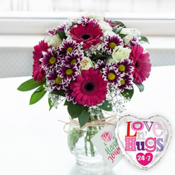 A lush, colour-rich flower arrangement stands in a clear glass vase, evoking the relaxed charm of Mill Hill's leafy streets. At the heart of the bouquet, striking gerbera daisies in strong magenta and fuchsia shades open in wide, confident discs, each with a deep, shadowy centre that anchors the composition. Surrounding them, clusters of chrysanthemums create a tapestry of jewel tones, their petals melting from velvety purple into pristine white at the tips, forming a crisp halo around warm yellow centres. Soft, creamy white roses are woven between these bolder hues, their unfurling petals adding a smooth, romantic texture that gently calms the intense colour play. Fine stems of baby's breath, studded with tiny white blossoms, weave in and out like a delicate lace, filling the spaces so the bouquet feels abundant yet airy. Fresh green foliage cradles and frames the flowers, the varied leaf shapes adding depth and a garden-like feel reminiscent of Mill Hill Park on a bright day. The transparent vase reveals the neatly trimmed stems, submerged in clear water and arranged in a careful spiral that hints at expert hands. Around the vase's neck, a simple length of natural twine is tied, supporting a small white tag adorned with a stylised pink rose and friendly green lettering, suggesting a hand-finished, artisan touch from Flowers Mill Hill. Slightly to the right, a shiny heart-shaped foil balloon with a silver border completes the scene, boldly printed with the words "Love n' Hugs 24-7" in a joyful mix of pink, purple and blue patterned letters. Resting on a light surface with a softly blurred, bright background, the entire setting radiates warmth and thoughtfulness, ideal for cheering someone in Mill Hill, from Anthony Road to the quieter residential lanes.