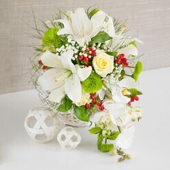 On a clean white tabletop that reflects a hint of its surroundings, a sophisticated floral arrangement in a round white wicker basket creates a serene focal point, as if prepared for an intimate wedding breakfast in Mill Hill. The background is a soft, textured beige fabric, gently lit so that no harsh shadows distract from the flowers. Rising from the basket, several large white lilies open with confidence, their trumpet-shaped petals showing fine speckles and deep throats dusted with golden pollen. They seem to lean slightly outward, suggesting a generous, open-hearted welcome. Nestled near the top among these lilies is a single cream-ivory rose, still fairly tight in its bloom, its petals arranged in a delicate spiral that adds warmth and subtle romance, reminiscent of a quiet stroll around Mill Hill Park in early spring. The bouquet is richly padded with mixed greenery: broader, glossy leaves close to the base, and slender, feathery foliage reaching outward, creating soft, sweeping lines. Tiny clusters of white baby's breath peek out between the leaves and petals, adding an airy, cloud-like texture. From the right side, a stream of white Phalaenopsis orchids cascades downward in an elegant arc, their smooth petals and nodding heads forming a gentle waterfall that gives the arrangement a graceful, elongated silhouette. Among the greens, bright green pom-pom flowers provide a modern, textured contrast, while clusters of small ruby-red berries add vivid points of colour, like tiny jewels hidden in the foliage. To the left of the arrangement, two decorative white spheres, crafted from thin interwoven strands, rest on the tabletop, echoing the basket's weave and giving a contemporary sculptural touch. The composition feels calm, refined, and celebratory, ideal for wedding receptions, anniversary dinners, or formal gatherings in and around Mill Hill.