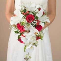 This enchanting scene from a Mill Hill bridal morning captures the serene moment as a bride, dressed in luminous white and delicate lace, clasps an elaborate cascading bouquet. Crafted with care by an expert florist, the bouquet bursts with pristine white lilies whose open faces invite gentle admiration, accented by sculptural stamens. Rich, ruby-red roses nestle closely among the lilies, their velvety textures symbolising the deep affection so often celebrated in Mill Hill marriages. Trailing phalaenopsis orchids flow from the main display, their graceful stems echoing the gentle slopes of Bittacy Hill and the tranquil elegance of local garden weddings. Scarlet berries-hypericum sprinkled with intention-dot the arrangement, offering vibrant, playful flashes of colour against clouds of soft white baby's breath. The supporting greenery, a lively mix of lacy fern and grounding leaves, seems to breathe fresh air into the floral composition, while the presence of spiky green accents injects hints of the untamed natural spaces found near Mill Hill Woods. The warm beige background blurs to nothing, turning all focus to the emotional beauty of the bouquet and the anticipation carried in the bride's arms. This bespoke arrangement celebrates Mill Hill's leafy charm, perfect for classic ceremonies in its historic chapels and picturesque venues.