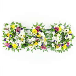 The photograph captures a classic "DAD" floral tribute, three large, freestanding letters resting in front of a plain white backdrop so that every bloom seems to glow. Each capital letter is carefully formed on a sturdy frame, its outline softened and enriched by a thick edging of lush green fern foliage. The fern fronds overlap and curl slightly, giving the letters a gentle, natural movement that contrasts with their bold, block shapes. Over this leafy foundation, the florist has layered a dense covering of fresh flowers until the surface feels almost velvety to the eye. White chrysanthemums and open white gerbera daisies dominate, their round, layered petals creating a calm and respectful base, reminiscent of quiet winter light over the greens of Mill Hill Park. Pockets of yellow - from bright gerberas to clusters of yellow spray roses - are woven through the white, adding warmth, hope, and hints of sunlight. Interspersed among these are smaller accents of rich purple lisianthus and vivid magenta or fuchsia spray roses, their deeper hues echoing the more complex emotions of grief and gratitude. Tiny lavender filler blooms and bright green button chrysanthemums nestle between the larger flowers, adding delicate detail and a lively touch of green. The three letters are linked subtly at the base by dark support bars, ensuring they read clearly as a single, loving message. This tribute feels personal and thoughtful, like something a family in Mill Hill might choose for a father's farewell, whether at a local church or crematorium, expressing love, pride, and remembrance through colour, texture, and care.