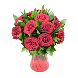 An opulent cluster of deep red roses, roughly twelve to fifteen blooms, rises in a compact, domed formation from a richly coloured red glass vase, positioned against a pure white background that allows the bouquet's intensity to take centre stage. Each rose is in that perfect stage of openness cherished by experienced florists: the petals are fully formed, spiralling outward in tight, velvety layers, yet still holding enough structure to promise several days of beauty. The roses' hue is a saturated crimson, reminiscent of red wine swirling in a glass, with soft highlights where the light brushes the petal edges and deeper maroon shadows tucked into the heart of each bloom. Threaded generously through and around these rose heads is a collar of fresh, dark green foliage, most likely ruscus, with smooth, slightly waxy leaves that arc gracefully and lend a natural, garden-like feel to the design. The foliage not only frames the roses but also adds contrast and volume, a deep emerald backdrop that makes the reds appear even richer. The vase itself, made of translucent red glass, has a gently narrowing waist and a flared rim, its subtly ribbed texture catching glints of light so that the glass looks almost like rippling silk. The stems inside are suggested rather than sharply defined, softened by the density of the colour. With its crisp, shadowless lighting and clean composition, the image suggests a modern floral studio in Mill Hill, perhaps just off Mill Hill Broadway, where a florist has created this piece as a sincere, no-words-needed gesture for anniversaries, intimate birthdays, or a passionate surprise delivery to homes and cafes stretching towards Mill Hill East.