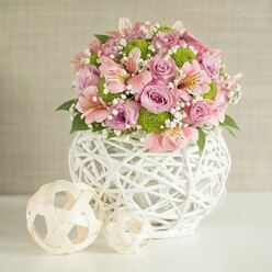 As I assemble this bouquet in our Mill Hill workshop, each element feels alive beneath my fingertips-the soft, velvety roses in pastel pink and lavender, the cool intricacy of a contemporary, woven white vase, and the lively greenery softly echoing the rolling landscape around Mill Hill Broadway. The composition radiates tranquility, its gentle blend of pinks and greens lightened by wisps of delicate baby's breath that glide between the blooms like a breeze through St. Joseph's College gardens. Among the roses, sprightly pink alstroemeria with streaked petals emerge, infusing the display with lasting charm, while crisp, lime green button chrysanthemums offer playful texture and a jolt of freshness reminiscent of springtime walks along The Ridgeway. Flanking the arrangement, two hand-woven decorative spheres in pale, natural tones provide visual continuity and balance, much like the harmonious streetscapes of Goodwyn Avenue or the artisan stalls found at Mill Hill Farmers' Market. Their placement-one large and sturdy closer to the vase, its smaller companion set forward-creates a sense of intentional artistry. The finished ensemble radiates subtle confidence and invites celebration, whether gracing a wedding table, a birthday lunch, or simply a bright spot in a Mill Hill family's home.
