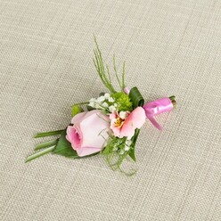 On a natural, pale beige linen reminiscent of Mill Hill's elegant tearooms, this buttonhole arrangement rests with quiet confidence, its composition a subtle dialogue between structure and spontaneity. The centrepiece-a blushing pink rosebud-spirals tightly, displaying a purity of form and hinting at the joy of fresh beginnings. To the side, a lighter pink or pastel peach flower, likely an alstroemeria or lisianthus, opens gently to reveal a touch of golden yellow and fine markings at its heart, the sort of botanical detail admired by floral lovers in Mill Hill's parks. Small, cloud-like tufts of baby's breath are tucked in, breaking up the palette with snowy elegance, as miniature cushion chrysanthemums in vibrant green add springtime energy. The foliage is arranged with the expertise of a seasoned Mill Hill florist: slender, verdant sprays shoot upwards while broader leaves at the base are precisely placed and at times folded for added depth. One larger green leaf wraps confidently around the stem's base, a modern touch mirroring the neat hedgerows along Mill Hill's byways. All is bound by a striking fuchsia satin bow-its rich sheen catching the light and heralding celebration. The boutonniere sits under soft, even lighting, every detail visible and each petal's gradation made to shine. Crafted for special moments-weddings, proms, or celebrations throughout Mill Hill-this floral pin carries with it the gentle aroma of garden mornings, the warmth of local tradition, and the timelessness that comes from authentic, heartfelt design.