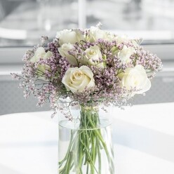 In this photograph, a meticulously arranged bouquet of creamy white roses and pale lavender statice rests in a tall, cylindrical glass vase on a smooth white tabletop, bathed in gentle daylight. The roses, likely a premium variety chosen for their symmetrical form and velvety petals, are fully developed yet not overblown, their outer petals curling back slightly to reveal warm, buttery centres. Their colour sits between soft ivory and antique cream, evoking a sense of timeless romance. Threaded among them are abundant tufts of tiny lilac statice blossoms, each branch creating a filigree of fine, papery flowers that surround the roses like a delicate mist. The statice adds depth and a subtle contrast of cool purple against the warmth of the white roses, making the bouquet feel both airy and full. Through the clear vase, you can see the cleanly trimmed stems, a fresh green that speaks of careful conditioning and same-day preparation, as you might expect from a trusted Mill Hill florist serving the quiet streets around Mill Hill Broadway. The stems are submerged in clear water, catching flecks of light and reflecting on the glossy surface beneath. The background is tastefully blurred, suggesting an interior space with a nearby window, allowing soft, natural light to wash over the flowers and give the petals a gentle glow. The mood is serene and composed, ideal for marking intimate moments: a small wedding celebration at a Mill Hill registry office, a thoughtful anniversary surprise, or a discreet sympathy arrangement. Every detail of the bouquet's round, balanced shape and limited colour palette emphasizes calm elegance and refined taste, capturing the quiet luxury that Flowers Mill Hill is known for across the local community.