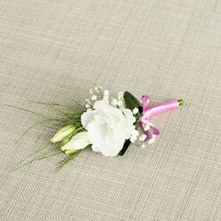 An elegant wedding boutonnière is displayed diagonally on a softly textured, oatmeal-toned fabric, reminiscent of fine linen often seen on reception tables in Mill Hill venues. At its centre sits a fully opened white lisianthus bloom, pristine and luminous, its many ruffled petals forming a rounded cup that looks both delicate and confident. Each petal curves and overlaps like fine silk, giving the flower a luxuriously soft, almost cloud-like presence against the warm neutral background. Nestled just to the lower left are two tall lisianthus buds, pale green with creamy hints at their tips, standing close together as if poised to open later in the celebration, echoing the sense of new beginnings that flows through wedding days along Mill Hill Broadway and surrounding residential streets. Sprays of white baby's breath weave through the composition, their tiny blossoms forming a misty, romantic frame around the central flower. Supporting this is a layer of rich, dark green foliage: a smooth, broad leaf anchors the design from behind, while slender, grassy fronds arc outward toward the bottom left corner, adding a sense of movement and a whisper of the garden. The stems at the base are tightly bound with a glossy fuchsia pink satin ribbon, tied into a neat, cheerful bow whose bright colour brings a playful, modern twist to the classic white and green palette. The gentle overhead lighting reveals every texture-from the fabric's weave to the satin's sheen-capturing a feeling of calm anticipation, as if this boutonnière is ready to be pinned to a groom's lapel just before he steps out for photos in one of Mill Hill's leafy side streets.