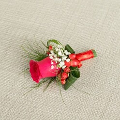 A handcrafted boutonniere rests on a pale beige, lightly woven fabric, reminiscent of a linen jacket or rustic wedding table runner you might see at a Mill Hill country-style celebration. The central bloom is a striking fuchsia-pink rose, its tightly layered petals spiralling outward in a rich, velvety swirl that suggests freshness and careful timing by the florist. Surrounding the rose, clusters of tiny white baby's breath create a soft, misty frame, their delicate blossoms offering a sense of purity and calm amid the brighter colours. Tucked between these white flowers are several plump red hypericum berries, their glossy skins catching the light and adding a cheerful, almost jewel-like sparkle. The greenery has been chosen and positioned with an expert eye: wispy fern-like fronds reach beyond the main cluster, adding movement and a touch of wildness, while darker, broader leaves are gently twisted and curled to form a sculpted collar around the rose, bringing structure and contrast. The stems are wrapped neatly in vibrant red satin ribbon, the fabric folded flat and smooth, with a neat bow or tuck giving the finish a tailored, comfortable feel against a suit lapel. Viewed from slightly above, the whole piece feels intimate and personal, like a quiet snapshot taken just before a groom steps out onto Daws Lane or towards a ceremony near Mill Hill Broadway. This boutonniere conveys romance, confidence, and celebration in a compact design, suitable for weddings, proms, or any formal event where a refined but colourful floral touch is desired.