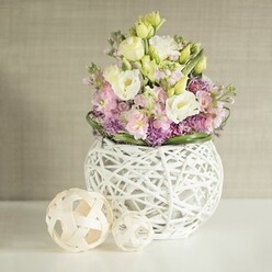 Bathed in gentle morning light that evokes the peaceful neighbourhoods of Mill Hill, a contemporary floral arrangement radiates quiet luxury and harmony. Within a stunning white spherical vase, crafted from intricately woven rattan, premium creamy lisianthus blooms mingle with delicate yellow petals, creating a sense of depth and softness. Pink stock flowers rise elegantly above the main cluster, their blooms unfurling with a gentle charm reminiscent of blooms seen in Mill Hill Park at the start of spring. A tufted base of plum and violet filler flowers-perhaps aster or miniature allium-adds a grounding richness and a textural contrast, all framed by an upturned band of deep green leaves that surround the bouquet. Resting gracefully to the left are two light rattan spheres, their openwork patterns subtly nodding to local crafts and home décor boutiques scattered along The Broadway. Set upon a bright white surface, with a softly textured cream wall behind, the arrangement feels warm, elegant, and lovingly curated. Every element, from bloom choice to composition, reflects the know-how and artistry of a florist rooted in Mill Hill tradition-an ideal centrepiece for wedding tables, family celebrations, or as a fresh spring accent in homes along Bittacy Hill.
