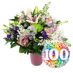 This image, set on a pure white background, captures the warmth and significance of a 100th celebration, like something you might see at a well-loved home off Mill Hill Broadway or near Mill Hill Park. Dominating the left side is a chic, ribbed vase finished in a soft matte pink, its subtle ridges adding texture without overwhelming the scene. From it rises a luxurious bouquet, densely arranged with fresh seasonal flowers that flow in a gentle dome of colour. Light pink lilies hold pride of place, some in full bloom with broad, star-shaped petals splayed open and their stamens reaching outward, others still enclosed in smooth buds reaching gracefully above the main cluster. Nestled around them are roses in varying shades of soft pink, mauve, and dusty rose, their layered petals forming plush, spiralled centres that draw the eye inward. Interwoven among the roses, rich purple lisianthus flowers introduce velvety depth, and small lavender asters with sunny yellow middles offer a more delicate note. Bright lime green button chrysanthemums provide playful bursts of texture, dotting the bouquet with compact, spherical blooms. Wisps of white baby's breath thread through the design, lending an airy, cloud-like softness, while mixed dark green foliage anchors everything with fullness and contrast, reminiscent of the mature trees lining local streets. To the lower right, slightly overlapping the flowers, a shiny round foil balloon adds an unmistakably festive touch. Its surface is alive with little confetti dots in a full rainbow of colours, and bold white numerals "100" appear centrally with a subtle 3D effect. The combination of elegant flowers and cheerful balloon perfectly illustrates a gift for a 100th birthday or 100-year anniversary celebration available for delivery throughout Mill Hill.