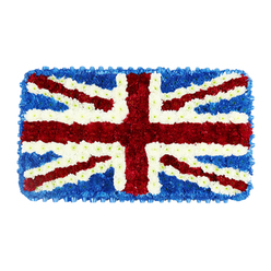 The image presents a striking floral Union Jack shaped as a neat rectangle, lying flat against a clean white background that allows every colour and contour to show with clarity. The entire flag is composed from countless small flowers set so close together they form a thick, cushion-like layer, evoking the softness of a well-worn tapestry rather than the stiffness of a fabric flag. A strong red St George's Cross dominates the centre, its vertical and horizontal lines formed from dense red blooms that create an unbroken sweep of colour. Around these red bands, crisply defined borders of white petals run alongside, tracing the familiar fimbriation that separates the red from the blue while keeping the overall design unified. From each corner of the rectangle, slender red diagonals of St Patrick's Cross travel towards the middle, again edged in white, lying over the broader white diagonal bands that represent St Andrew's Cross. The remaining fields and the perimeter are filled with deep blue florets that might remind a Mill Hill florist of tightly clustered hydrangeas, creating a steady, dignified backdrop. The texture is richly three-dimensional, with each flower head slightly raised, catching light in subtle highlights and shadows. This floral tribute feels particularly moving as a funeral or memorial piece for someone connected to British traditions, perhaps remembered after years in the Mill Hill community near the Broadway, local synagogues, or the green spaces around Arrandene. It radiates a mood of shared memory and quiet pride, with the careful craftsmanship reflecting the respect and care given to the person being honoured.