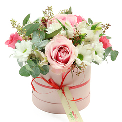 Centered against a pure white background, a pastel pink hatbox brims with flowers, giving the impression of a special gift just arranged on a Mill Hill kitchen table in the soft light of morning. The cylindrical box has a smooth, matte finish and is encircled by a coral-red satin ribbon, its sheen catching the light as it is tied into a tidy bow at the front. From the bow hangs a rectangular cream gift tag with flowing green handwriting, evoking a personal note or dedication just before the bouquet is dispatched to a home near Mill Hill Broadway or a quiet side street off Daws Lane. Above this elegant base, the bouquet rises in a composed, domed form. A striking rose, pale pink around the edges with a more intense pink swirl at the centre, claims pride of place, its spiralled petals layered like soft silk. Surrounding it are pure white daisy-like chrysanthemums with fresh green centres, their bright faces bringing clarity and light to the arrangement. White alstroemeria, distinguished by their slim, trumpet-shaped petals and delicate inner speckles, weave through the design, softening transitions between the larger blooms. Among them, clusters of smaller, rich fuchsia or deep pink flowers-likely spray roses or lisianthus-provide vibrant accents that suggest romance and warmth. The entire composition is threaded with foliage: rounded, silver-green eucalyptus leaves that introduce a cool, contemporary note, and deeper green leaves that anchor the colour palette. Here and there, fine stems tipped with tiny buds add a natural, almost wild touch, as though clipped from a garden in nearby Mill Hill Park. The overall mood is one of thoughtful elegance and comforting beauty, perfect for same-day flower delivery in Mill Hill when words alone do not feel enough.