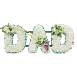This image portrays a classic "DAD" funeral tribute, the three letters crafted with the careful precision of an experienced Mill Hill florist. Each letter stands upright and is fully covered in pristine white chrysanthemums, the small, round blooms packed tightly together to create an even, cushion-soft texture. The effect is of crisp, snowy blocks of flowers, the petals forming a smooth, continuous surface that reads clearly even from the back of a chapel or along a roadside during a funeral procession. Around the outer edge of every letter runs a band of deep emerald green ribbon, gently ruffled and slightly glossy, giving a smart, tailored finish and framing the white flowers with a natural, verdant touch. Behind the letters, subtle glimpses of a dark supporting frame can be seen, revealing the structure that keeps the tribute secure and neatly aligned.   

Delicate floral accents bring movement and personality to the design. On the upper left side of the first "D", a small spray of white freesia blooms and polished greenery is tucked into the flowers, the freesia buds and trumpets adding texture and a light, sweet fragrance. Two long green stems curve gracefully from this point towards the central "A", creating a soft diagonal line that links the letters. At the lower right of the "A", where the stems meet, a more dramatic spray becomes the focal point: two deep purple calla lilies, their sleek, rolled trumpets rich and velvety, rest among additional white freesia and bright foliage. The final "D" is topped on its upper right curve with another, slightly smaller cluster of white freesia and leaves, mirroring the first letter and giving the arrangement pleasing balance.   

Set against a clean, white background, the tribute radiates a sense of calm respect and quiet sorrow. With its restrained colour palette and elegant detailing, it feels particularly fitting for a Mill Hill farewell, whether placed in a local church, a funeral home near Mill Hill Broadway, or carried past the open green spaces that families in the area know so well.