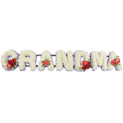 In this tender sympathy arrangement, the word "GRANDMA" is spelled in large, three-dimensional letters crafted entirely from fresh white blooms, evoking the quiet dignity of a funeral service in Mill Hill or a memorial gathering at a nearby church. Each capital letter is meticulously formed from densely packed white cushion chrysanthemums or similar small-headed flowers, their petals overlapping to create a smooth, plush texture that reads as soft and comforting. The outline of every letter is gently defined by a narrow border of pale purple ribbon or backing, which peeks out like a soft lilac shadow along the curves and edges and helps each character stand out clearly against the bright white background. Vivid red accents are arranged with purpose and care. On the first letter, "G", a small spray of deep crimson roses is nestled neatly into the lower curve, alongside an elegant red-and-white variegated calla lily whose curved funnel shape adds a sculptural feel. The initial "A" is highlighted by a single, heart-shaped red anthurium at its base, its smooth, lacquered surface almost glowing. The central "M" is crowned with another grouping of red roses and a matching red anthurium, creating a focal point at the heart of the tribute, while the final "A" repeats the motif with one more solitary red anthurium. A dark green stand or frame discreetly supports the entire word, giving the impression of stability and continuity when seen from the front. The overall mood is one of reverence, warm memories, and gentle farewell, as though a family in Mill Hill has chosen a floral tribute that speaks their love for a much-adored grandmother without needing any words spoken aloud.