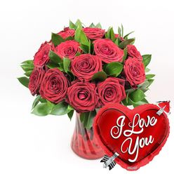 In this evocative floral image, a sumptuous bouquet of deep red roses is the undeniable focal point, framed by lush greenery and accompanied by a gleaming heart balloon that together create a scene of classic romance. The bouquet, containing approximately a dozen or more roses, is tightly arranged into a rounded, full shape, each bloom angled to show off its velvety petals. Some roses are fully open, with layers of petals spiralling toward the centre like tiny crimson whirlpools, while others remain partially closed, suggesting unfolding devotion over days to come. Nestled among the roses, rich green foliage peeks through, its leaves smooth and slightly glossy, adding both volume and contrast so the red tones feel even more intense. These stems are placed in a translucent ruby-red glass vase with elegant fluted sides; the vertical ridges create subtle shadows and reflections that give the impression of fine craftsmanship you might expect from an experienced Mill Hill florist. Clear water inside the vase anchors the stems and hints at their freshness, as though the arrangement has just been prepared for delivery across Mill Hill and Totteridge. In the lower right corner of the image, overlapping the vase and part of the bouquet, a bright red heart-shaped foil balloon introduces a playful, celebratory element. Its metallic surface glows with reflected light, showing gentle contours and a soft sheen. Across its centre, in bold yet graceful white cursive, the words "I Love You" are written clearly. A stylised silver arrow pierces the heart diagonally, from lower left to upper right, echoing the beloved symbol of Cupid. Set against a crisp white background, the rich reds, vibrant greens, and reflective metallics are accentuated, creating a powerful visual statement of affection perfect for Valentine's Day surprises, anniversaries in Mill Hill, or any romantic occasion where words alone feel too small.