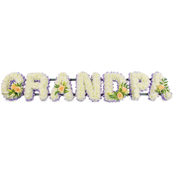 The image shows an elegant "GRANDPA" funeral tribute, arranged as a long row of freestanding floral letters that seem ready to be placed beside a coffin or at the front of a service in Mill Hill. Each letter-G, R, A, N, D, P, and the final A-is completely filled with tightly packed white chrysanthemums or similar small, daisy-like blooms. The flowers sit closely together, forming a plush, cloud-soft surface where their tiny yellow-green centres are barely visible, lending the piece a calm, pure look. Around the perimeter of every letter, a narrow ring of soft lavender or lilac flowers traces the outline, adding a gentle wash of colour and sharpening the silhouettes without overpowering the serene white. Thoughtful accent sprays add warmth and individuality: on the "G" and "D", clusters of peach-toned roses, some fully open and others just starting to unfold, sit among slim white lily buds and rich green foliage, bringing a natural, garden-like touch to the tribute. Single peach roses with greenery decorate the first and last "A", echoing these larger clusters and guiding the eye along the word. All the letters rest on a slim, dark support bar that runs beneath them, offering structure and stability while remaining largely hidden. Photographed against a clean white background, the textures and colours appear crisp and defined, reminiscent of walking into a quiet chapel off Mill Hill Broadway where a carefully prepared arrangement waits in stillness. The tribute gently expresses love, gratitude, and remembrance for a grandfather who meant the world to his family.