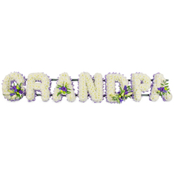 In this image, a lovingly crafted floral lettering arrangement spells out the word GRANDPA in bold, individual letters, arranged along a single horizontal line as if ready for display at a funeral or memorial service in Mill Hill. Each letter is richly covered with small white chrysanthemums or carnations, the flowers so densely packed that they form a uniform, velvety surface without visible gaps. The petals create a soft, textured effect, like a series of white cushions, conveying purity, peace, and the gentle comfort families seek when saying goodbye. Subtle yet meaningful accents of deep purple and lavender flowers are threaded through the white letters, possibly lisianthus or miniature chrysanthemums, accompanied by fresh green foliage. These accents are placed thoughtfully: a cluster graces the base of the G, another rests within the hollow of the first A, more blooms appear in the body of the D, then along the stem and curve of the P, and finally near the last A, adding points of interest and guiding the gaze along the tribute. The green leaves peeking out between petals introduce a natural freshness that enhances the solemn beauty of the design. Underneath, a dark green bar or frame runs discreetly from one end of the word to the other, providing essential support but remaining largely hidden by the blooms. The plain, light background allows the form and colour of the tribute to stand out clearly. This type of bespoke memorial piece, often arranged by local florists serving Mill Hill, Totteridge, and nearby chapels, offers a deeply personal way for a family to honour their grandfather's life, turning their affection and memories into a dignified floral message.