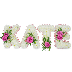This luminous, handcrafted name tribute reads KATE in substantial three-dimensional letters, each letter built from densely packed, pristine white chrysanthemums that form a seamless, cloud-like surface. The chrysanthemums are arranged with precision so the edges are crisp yet soft to the eye, and a pale pink satin ribbon delicately outlines the internal shapes, adding a refined, feminine frame that catches morning or chapel lighting and imparts a faint sheen. Hot pink roses serve as focal punctuation across the name: a double cluster on the K, a single, perfectly open rose nestled low in the A, a solitary rose grounding the T, and twin roses brightening the E-each bloom tucked into swathes of deep and light green foliage, with subtle touches of tiny pink buds and alstroemeria for texture. Ferny fronds and glossy ruscus or eucalyptus leaves braid through the composition, lending movement and an herbaceous scent beneath the sweeter rose perfume. The letters are freestanding and cast gentle shadows on the white surface below, creating a three-dimensional presence suited to memorial services, graveside tributes or a quiet celebration of life. As a local Mill Hill arrangement, it reads as both intimate and ceremonious-thoughtfully personalised, crafted to be both visually clear from the front row and tender up close, offering a comforting visual of love and remembrance for family and friends.