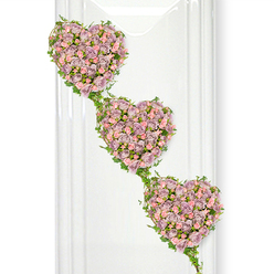 Three interlinked hearts made entirely of roses form the centrepiece of this serene funeral arrangement, their shapes descending diagonally as though they are gently falling into place. Each heart is built from tightly clustered miniature roses in muted mauve, dusty lavender, and soft pastel pink, the colours blending together with a subtle, romantic softness. The roses are arranged so densely that they create a rich, velvety surface, with some blooms fully opened to show their layered centres while others remain as rounded buds, hinting at new life within the tribute. Framing and connecting the hearts are trails of ivy, their slender stems looping in graceful curves and their small, glossy green leaves adding structure and contrast. The ivy clearly outlines each heart before it travels on to the next, forming a natural chain that speaks of continuity. The background is bright white, with delicate vertical reflections to one side, suggesting a minimalist, polished setting-perhaps reminiscent of a light-filled room in Mill Hill or the calm interior of a local chapel off Flower Lane or Bittacy Hill. The lighting is soft, almost diffused, allowing every petal and leaf to be seen without distraction. The overall impression is one of quiet, dignified affection, making this design deeply suited to a memorial or funeral where the family wishes to express an unbroken bond. It is easy to imagine this triple-heart tribute resting by the casket or beside a framed photograph, a handcrafted expression of love that a Mill Hill florist has arranged with particular care for the final goodbye.