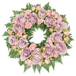 This image shows a beautifully crafted circular wreath laid flat on a pure white surface, its design inviting a long, thoughtful look. The wreath is densely filled with roses, the dominant blooms being large, fully open flowers in a sophisticated palette of dusty lavender, muted mauve, and cool, greyish purple. Their many layered petals curl gently at the edges, giving a soft, ruffled texture that repeats around the ring. Tucked between these statement flowers are clusters of smaller blush pink spray roses and scattered buds in different stages of opening, which introduce variety and a sense of gentle progression, like memories surfacing over time. Underpinning the design is a deep, textured layer of dark green fern fronds, their fine, divided leaves creating a feathery cushion that stretches slightly beyond the outer line of blooms. Here and there, darker filler greenery and tiny, nearly closed buds are woven through the roses, adding dimension and subtle contrast without disturbing the wreath's serene harmony. The colour combination of lavender, pink, and rich green creates a calm, consoling mood, suited to moments of reflection and farewell. It is easy to imagine this wreath being delivered to a funeral or celebration of life service in Mill Hill, perhaps placed near the front of a chapel or at a graveside after a quiet walk from Mill Hill Broadway. The soft, even lighting highlights the natural textures of petal and leaf, underscoring the care and intention behind each placement-a floral expression of sympathy and enduring love from a trusted local florist.
