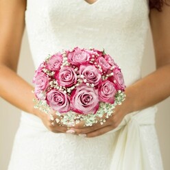 In a moment that radiates the serene elegance of a Mill Hill celebration, this close-up encapsulates the anticipation and artistry of a bride's most cherished bouquet. The compact, hand-tied arrangement, designed by skilled florists who know the NW7 area intimately, glows with nearly a dozen roses in gradient hues-from soft baby pink at the inner petals to vivid magenta ringing the edges. Each bloom is at its peak, spread open in perfect spirals, while fine sprays of baby's breath cocoon the roses, adding a feathery, cloud-soft dimension reminiscent of a spring breeze in Mill Hill Park. The bride's arms, tanned by gentle sunlight, and immaculately manicured hands cradle the bouquet with a tenderness that speaks of new beginnings. Her dress-a sleeveless vision in white floral lace with an elegant V-neckline-is gathered gracefully with a sash to the side, nodding to the refined tastes found in the area's wedding circle. Hints of greenery frame the bouquet's crown, grounding the composition in natural beauty. Set against a subtle, neutral background, the arrangement becomes the focal point, a timeless symbol of hope and joy. Every detail-from the lacework to each petal and wisp-embodies the spirit of Mill Hill's most memorable occasions, inviting couples searching for inspiration in local venues like Mill Hill Synagogue or Copthall Fields to imagine these blooms as part of their own love story.