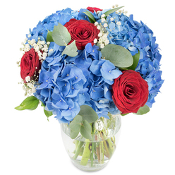 Displayed against a pristine white background, this hand-crafted bouquet rests in a transparent glass vase with a gently rounded base and a tapered top, the green stems clearly visible through fresh water. Broad clusters of vivid sky-blue hydrangeas form the plush foundation, their many small florets overlapping into a soft, cloud-like hemisphere that anchors the design. Contrasting with this cool expanse are several intensely red roses - their velvety, tightly coiled petals offering concentrated spots of romantic colour that draw the eye. Wisps of white baby's breath are delicately interspersed, lending a light, celestial texture and a sense of movement among the larger blooms. Soft, rounded eucalyptus leaves with a muted, sage-green tone are threaded through the bouquet, providing sculptural shape and a subtle herbal fragrance that lifts the arrangement. Bright, even lighting highlights the saturated blues and reds and emphasizes the crisp freshness of the stems in water. As a local Mill Hill florist I've placed arrangements like this for tables at Mill Hill Broadway gatherings and village celebrations; it reads as both a refined centerpiece and a heartfelt gift for anniversaries, patriotic moments, or to mark life's milestones. Flowers Mill Hill prefers this classic trio of colours when a customer asks for something bold yet composed - a bouquet that feels both sophisticated and warmly familiar to neighbours across the area.
