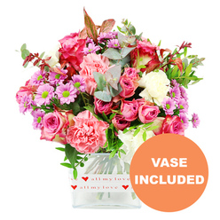 In this photograph, a generous, dome-shaped bouquet fills a clear, square glass vase, standing out vividly against a pure white backdrop so every flower and leaf is clearly visible. The arrangement leans into a romantic palette of pinks and soft pastels: velvety roses in deep fuchsia and gentle blush tones open in layered spirals, some fully in bloom and others still slightly cupped, suggesting freshness. Nestled among them are lush carnations with their signature ruffled edges, in shades of coral, peach-pink, and creamy white that add both volume and a sense of softness, like a cosy throw over a chair in a Mill Hill sitting room. Dotted between the larger blooms are small daisy-like chrysanthemums in lavender and light purple, each flower centred with a bright green eye that injects a lively, sparkling contrast. The bouquet is supported by a collar of rich greenery, including rounded eucalyptus leaves in cool blue-green, deeper green foliage, and the occasional reddish-brown leaf that introduces a subtle warmth reminiscent of autumn walks around Arrandene Open Space. The clear glass vase, slightly rounded at the corners, is wrapped around its lower portion with a white band printed with tiny red hearts and the phrase all my love in an elegant script, instantly marking this as a bouquet meant to convey emotion-be it romantic love, thanks, or encouragement. In the lower right-hand corner of the picture, a bold orange circle with white capital letters reads VASE INCLUDED, making it clear that the bouquet arrives complete and ready to place on a table in a Mill Hill kitchen, office, or reception area, perfect for thoughtful same-day deliveries across the neighbourhood.