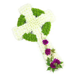 In this serene studio image, a floral Celtic cross lies against a clean, bright white backdrop, its traditional shape clearly defined by an intricate combination of white and green blooms. The main structure of the cross-its vertical stem and extended arms-is made from tightly packed white chrysanthemums, each small head forming part of a dense, cushion-like layer that appears soft yet reassuringly solid. Surrounding and threading through this white canvas, lime green button chrysanthemums have been precisely arranged to draw out the hallmark features of a Celtic cross. They form a striking circular halo where the vertical and horizontal arms meet and trace gentle knot-like patterns that add movement and symbolism, suggesting heritage and continuity. At the lower part of the cross, three prominent clusters of deep magenta-purple carnations create a focal point, their frilled, velvety petals rich with colour and emotion. Fine strands of dark green foliage weave delicately around these purple blooms, some of them curling like ivy, softening the outlines and adding a touch of natural grace. The lighting is even and clear, revealing the freshness of the flowers and the florist's careful craftsmanship. This arrangement would feel at home in a quiet chapel service in Mill Hill or placed lovingly beside a memorial at Mill Hill Cemetery, where families gather to say goodbye. The blend of white, green, and purple radiates calm, respect, and hope, reflecting the thoughtful work of local florists who understand how to create tributes that speak gently yet powerfully of remembrance and faith.