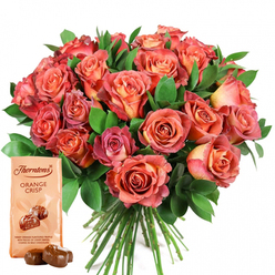 There's a refined joy in this hand-crafted arrangement, as if it had been gathered by a Mill Hill florist with years of experience spreading beauty through NW7. The main attraction is a lavish, rounded bouquet filled with roses in every warm hue-gentle coral, tender peach, lively salmon, and saturated rosé pinks. Each rose appears brushed with nature's own ombre, some featuring soft yellow or cream at the centre and deepening to fiery pinks at the tips-an echo of Mill Hill's summer sunsets. The flowers are nestled within a generous arrangement of dark, glossy green foliage, providing a cool counterpoint and accentuating the abundance that characterises the bouquet. At the base, to the left, an upright Thorntons Orange Crisp chocolate pouch subtly invites with its light cream wrapper, signature branding, and a tantalising description illustrated with glossy, round truffles. Two real truffles lean temptingly before the pouch, their smooth milk chocolate coating shimmering under soft lighting. The entirety feels composed not only for visual delight but to awaken the senses and create memories-making it a natural choice for marking new beginnings, special birthdays, or anniversary milestones in Mill Hill. The artistry and heart shine through in every detail, a gesture as comforting as it is lavish.
