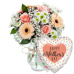 This image shows a tender, celebratory Mother's Day arrangement styled with the quiet elegance you might expect from an experienced Mill Hill florist. Against a spotless white background, a full mixed bouquet is presented in a clear glass vase, its shape simple and rounded so that the focus stays on the flowers. Around the neck of the vase, a loop of light brown twine is tied into a small bow, adding a rustic accent that makes the arrangement feel personal and homemade, like a lovingly prepared gift waiting on a dining table near Mill Hill Park. The bouquet features several beautifully open roses in shades of peach and blush pink, their petals soft and velvety, arranged in spirals that invite the eye to linger. Nestled close by are broad-faced peach gerbera daisies, each one centred with a rich, dark brown disc that grounds the pastel tones and adds visual contrast. Between the larger blooms, delicate sprays of white baby's breath drift through the arrangement, forming airy layers that echo fine lace. Small, rounded white chrysanthemums with gentle green centres increase the sense of fullness and contribute a crisp, clean brightness. Sprigs of verdant foliage weave between the flowers, offering depth and a hint of the natural, leafy scent that accompanies freshly cut stems. Behind the blooms, partially hidden, a small white greeting card peeks out, its coloured writing suggesting a handwritten "Happy Mother's Day" from a son or daughter. In front of the vase, leaning slightly to the right, a large heart-shaped foil balloon acts as a playful counterpoint to the flowers. Its white background is dotted with metallic gold and silver spots, like tiny confetti pieces scattered across the surface. At the centre, a softly textured peach heart frames the message "HAPPY Mother's DAY" rendered in stylish black type, with "Mother's" in flowing calligraphy that adds a touch of sophistication. The overall composition radiates joy and appreciation, making it a fitting visual for Mother's Day flower delivery in Mill Hill, whether to homes along Hale Lane or townhouses near Mill Hill Broadway Station.