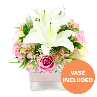 A delicately balanced pink and white floral arrangement rests against a plain white background, its soft colours and precise structure suggesting the handiwork of a skilled local florist in Mill Hill. Dominating the centre are striking white Oriental lilies, their petals broad and gently recurved, forming a sculptural star shape that draws the eye in to the dark reddish-brown stamens standing proudly in each bloom. Around these lilies, numerous soft pink roses are nestled, some still in tight buds with smooth, spiralling centres, others fully open with many layers of petals that create a lush, cushion-like texture. Threaded through the roses are stems of alstroemeria in gentle shades of blush and ivory, their slim petals enhanced by light yellow tones and fine speckles, adding delicate visual interest. Wispy sprays of white baby's breath are scattered throughout, introducing a sense of lightness and space, as though a soft breeze from Mill Hill Park were passing gently through the bouquet. Rich green foliage surrounds the flowers and peeks between them: shiny, broad leaves form a solid base, while lighter, feathery greens add movement and a fresh, garden-picked character. The entire composition is arranged in a modern, square clear glass vase, with the neatly trimmed stems visible through the water, emphasising the freshness of the design. A pale pink satin ribbon is wrapped horizontally around the middle of the vase, providing a subtle decorative touch without distracting from the blossoms. In the lower right corner of the image, a bright orange circular label with bold white capital text announces VASE INCLUDED, making it easy to see that the container comes with the flowers. This bouquet feels serene yet celebratory, ideal for a bedside table in a Mill Hill home or as a charming gift delivered to mark birthdays, thank-yous, or new beginnings.