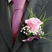 The image presents an intimate, waist-up view of formal attire, capturing the quiet drama of a single boutonniere pinned to a dark charcoal or black suit jacket, as if the wearer is about to step into a wedding venue in Mill Hill. A crisp, pale dress shirt with a fine gingham or checked pattern peeks out from beneath the lapels, its subtle texture playing gently against the smooth, satiny sheen of a vivid fuchsia tie that runs straight down the centre of the chest. Anchored to the left lapel, the boutonniere is centred on a perfectly opened light pink rose, its petals soft and velvety, graduating from a more intense rose-pink at the base to a delicate blush at the tips. Around this focal bloom, airy sprigs of white baby's breath cluster in tiny, starry blossoms, giving the arrangement a cloud-soft halo that contrasts with the sharp tailoring of the jacket. Fine strands of bright green foliage lift and fan out behind the rose, adding height, freshness, and a touch of wild garden character that feels reminiscent of a walk through the greenery near Arrandene Open Space. At the base, a broader, glossy green leaf is wrapped snugly around the stems, secured with a neat purple ribbon tied in a small, carefully formed bow that sits gracefully above the suit's pocket line. The soft lighting reveals every contour of petal and thread of fabric, suggesting a calm, hopeful moment just before vows, prom photos, or a gala reception. The composition captures the blend of formality and emotion that local florists in Mill Hill create for life's milestone celebrations.