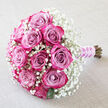 Set on a gentle diagonal over a pale, textured fabric, this bouquet looks ready to be lifted by a bride just outside a church in Mill Hill. A compact cluster of around twelve roses takes centre stage, each bloom displaying a beautiful colour shift from soft, dusky mauve and lavender-pink in the middle to bright fuchsia and deep magenta along the outer petals. The roses vary between tightly closed and softly opening, so that the spirals of velvety petals create layers of depth and a feeling of quiet anticipation. Embracing these vibrant blooms is a full collar of white baby's breath, with countless Gypsophila florets forming a fine, airy mist around the roses. This cloud of white gently diffuses the colour, creating an almost ethereal border that recalls the lightness of a bridal veil or the soft haze of a summer morning in Mill Hill's open spaces. Here and there, discreet touches of dark green leaves appear, anchoring the arrangement with a natural, garden-inspired note. The stems are neatly bundled at the lower right and carefully wrapped in a lilac satin ribbon, its smooth, slightly lustrous surface reflecting light in a calm, elegant way. Coiled delicately around the ribbon is a slim strand of white pearls, each bead placed to suggest hand-finished craftsmanship and traditional celebration. The bouquet is photographed under soft, even light that reveals each curve and texture, making this design ideal for weddings, vow renewals, or romantic gestures delivered to homes and venues throughout Mill Hill and the surrounding area.