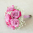 This photograph shows a petite yet luxurious hand-tied bouquet placed on a neutral, light beige fabric surface with a gentle woven texture, allowing every floral detail to stand out. At the heart of the bouquet is a tight dome of exquisite roses, each one displaying a beautiful two-tone colour play. Their inner petals are washed in cool lavender and soft purple hues, while the outer edges are brushed with vivid magenta and rich fuchsia pink, creating a layered effect that suggests depth and movement. Interlaced between these focal blooms is a generous mist of white baby's breath, the fine gypsophila florets forming airy clusters that soften the strong rose colours and give the design a classic, romantic feel. The flowers are gathered into a short, tidy handle wrapped in deep purple satin ribbon, its surface smooth and slightly glossy. Wound gracefully around this ribbon is a string of faux white pearls, spiralling down the handle like a delicate bracelet and adding a subtle touch of bridal elegance. The bouquet lies at a diagonal angle, with the pearl-decorated stems pointing towards the right-hand side of the image, suggesting it could be lifted at any moment for wedding photos near Mill Hill Broadway or carried down the aisle of a local chapel. The soft lighting creates minimal shadows, highlighting the intricate petal edges and the contrast between the vibrant roses, the crisp white gypsophila, and the calm background. Together, these elements evoke a sense of intimate celebration, perfect for weddings, prom nights, or engagement parties in and around Mill Hill.