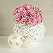 In this composition, a neat dome of pink roses rises from a white woven orb, framed by a soft collar of baby's breath, creating a floral sculpture that feels perfectly suited to a Mill Hill wedding. The roses are densely packed, their velvety petals overlapping like layers of silk. Each blossom transitions gently from a pale, cool pink at its heart to a richer, almost berry-like shade at the petal edges, adding depth and a painterly quality to the bouquet. Nestled between the roses and gently skimming the outer edge, delicate stems of gypsophila provide a cloud of tiny white blooms, giving the design a light, airy aura, as though the roses are floating on a cushion of snow. The flowers are set in a spherical, hand-woven vessel made from slim white strands that criss-cross and curl around each other, forming an open globe. The empty spaces between the weaving let the surrounding light in, throwing subtle, looping shadows on the tabletop and enhancing the sense of movement and space. The overall atmosphere is calm and joyful, reminiscent of a wedding breakfast near Mill Hill East, where clean lines and soft colours bring a sense of quiet luxury. You can almost imagine this piece placed along a line of tables in a bright marquee, the scent of rose drifting gently as guests raise their glasses. The arrangement pairs especially well with contemporary venues around Mill Hill Broadway and Totteridge, where couples look for designs that are simple, elegant, and full of feeling. It would suit engagement dinners, bridal showers, or romantic anniversary celebrations, bringing a modern touch while still honouring the timeless beauty of pink roses.