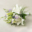 Bathed in soft, natural light on a neutral beige fabric, this elegant bouquet centres on pristine white lilies whose wide, satiny petals curl back to reveal golden anthers and tiny dark speckles within their throats. Beside them, lisianthus add a painterly touch: white blooms edged with a delicate lavender border and ruffled centres that move between tight buds and open corollas, creating an inviting rhythm of forms. Wisps of feathery green foliage thread through the cluster, pairing with broader deep-green leaves to give the bouquet a grounded fullness, while small sprays of baby's breath lift the design with an airy, cloud-like texture.

The gathered stems are tied neatly with a white satin ribbon at the right, the glossy wrap reflecting the light and signalling a careful hand in the arrangement's construction. The linen backdrop and the soft shadows suggest a sunlit table in a quiet Mill Hill home, evoking early-summer calm and subtle celebration. This arrangement reads as refined and approachable - quiet enough for a sympathy or bedside bouquet, yet composed enough for a wedding table or thoughtful gift. Its balance of texture, scent, and soft colour makes it an invitation to pause and savour a gentle, floral moment.