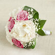 On a softly textured, off-white linen surface, this hand-tied bouquet is displayed as though resting between photos at a wedding in Mill Hill. At the heart of the arrangement lies a single, generous creamy white peony, fully open and lush, with layers of finely ruffled petals that seem to billow outward like soft fabric. Encircling this focal bloom are several roses in vivid fuchsia-pink and muted blush, their spiralled petals tightly layered and luminous in the gentle light, adding both drama and warmth to the design. Threaded through the arrangement, delicate heads of white gypsophila, commonly known as baby's breath, bring a lace-like texture, their tiny blossoms creating a misty, romantic veil over the stronger colours. Broad, dark green leaves cradle the bouquet from below and around the sides, their smooth, glossy surfaces arching gracefully and providing structure, reminiscent of the well-kept gardens and tree-lined streets around Mill Hill Broadway. The stems are neatly gathered and secured with a band of pure white satin ribbon, wrapped snugly to form a sleek, practical handle, suggesting that this bouquet is made to be carried comfortably through a full day of celebration. Gentle, even lighting enhances the creamy whites and rich pinks, while the linen background remains neutral and understated, allowing the flowers to take centre stage. This arrangement conveys a mood of serene romance and polished charm, suitable for bridal parties, anniversary surprises, or special deliveries to homes and venues throughout Mill Hill and nearby North London areas.