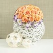 A carefully composed centerpiece sits on a smooth white surface, bathed in soft light that gently highlights every curve and petal, as though prepared for a wedding table in Mill Hill or a stylish gathering near Mill Hill Broadway. At the centre is a dome of roses in tender peach and apricot hues, the blooms tightly grouped to form a near-perfect sphere of velvety petals. Each rose is open and generous, with layered spirals that overlap and nestle together, creating a plush, seamless appearance that radiates warmth. Just below this rose dome, a delicate collar of lisianthus unfolds, their white blossoms finely frilled and edged in deep violet or purple, adding a crisp, refined outline that encircles the warmer tones above. Between them, small clusters of white baby's breath emerge, each tiny flower like a soft dot of light, lending airiness and a sense of movement to the otherwise smooth silhouette. The flowers are held in a distinctive, broadly rounded container crafted from white woven wicker or rattan, its crisscrossing strands forming an open lattice that adds texture and a contemporary, hand-crafted feel. Light filters through the gaps, casting subtle shadows that give depth to the design, the way afternoon light plays across the greenery in nearby Mill Hill Park. In front of the container, two decorative spheres rest on the tabletop-one larger, one smaller-made from pale wooden strips woven into open, nest-like orbs that visually echo the vase's structure and add a sculptural touch. Behind it all stretches a softly textured beige backdrop, reminiscent of natural linen or burlap, providing a warm, neutral canvas that lets the flowers' gentle colours shine. The mood is serene, romantic, and thoughtfully elegant, suited to weddings, anniversaries, or intimate celebrations throughout Mill Hill.
